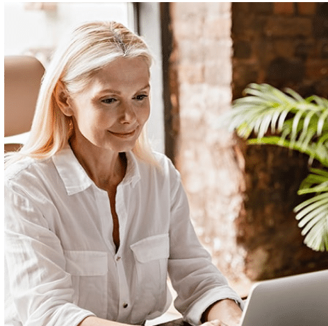 Woman with blonde hair working on laptop in a cozy home office with a green plant.