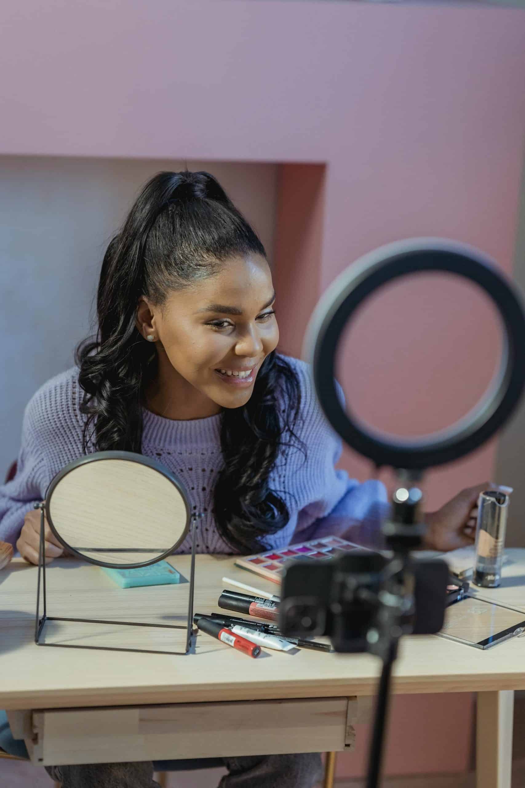 Cheerful makeup creator smiles at a stylish desk filled with beauty products under bright lighting.