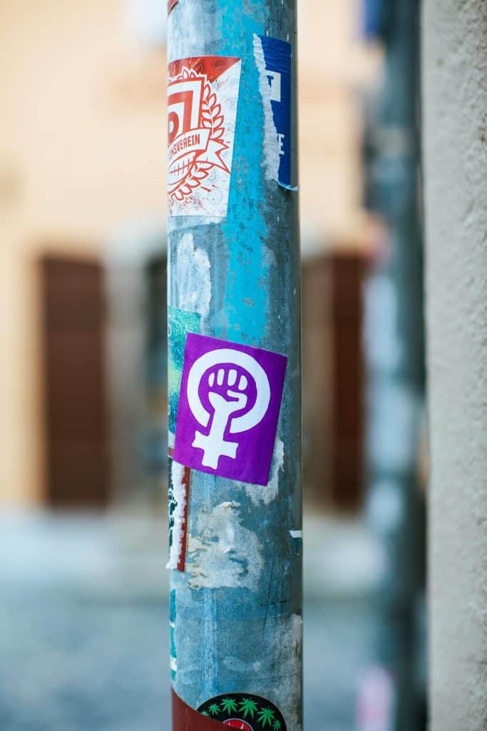 Weathered pole adorned with vibrant stickers, featuring a striking purple feminist symbol of empowerment.