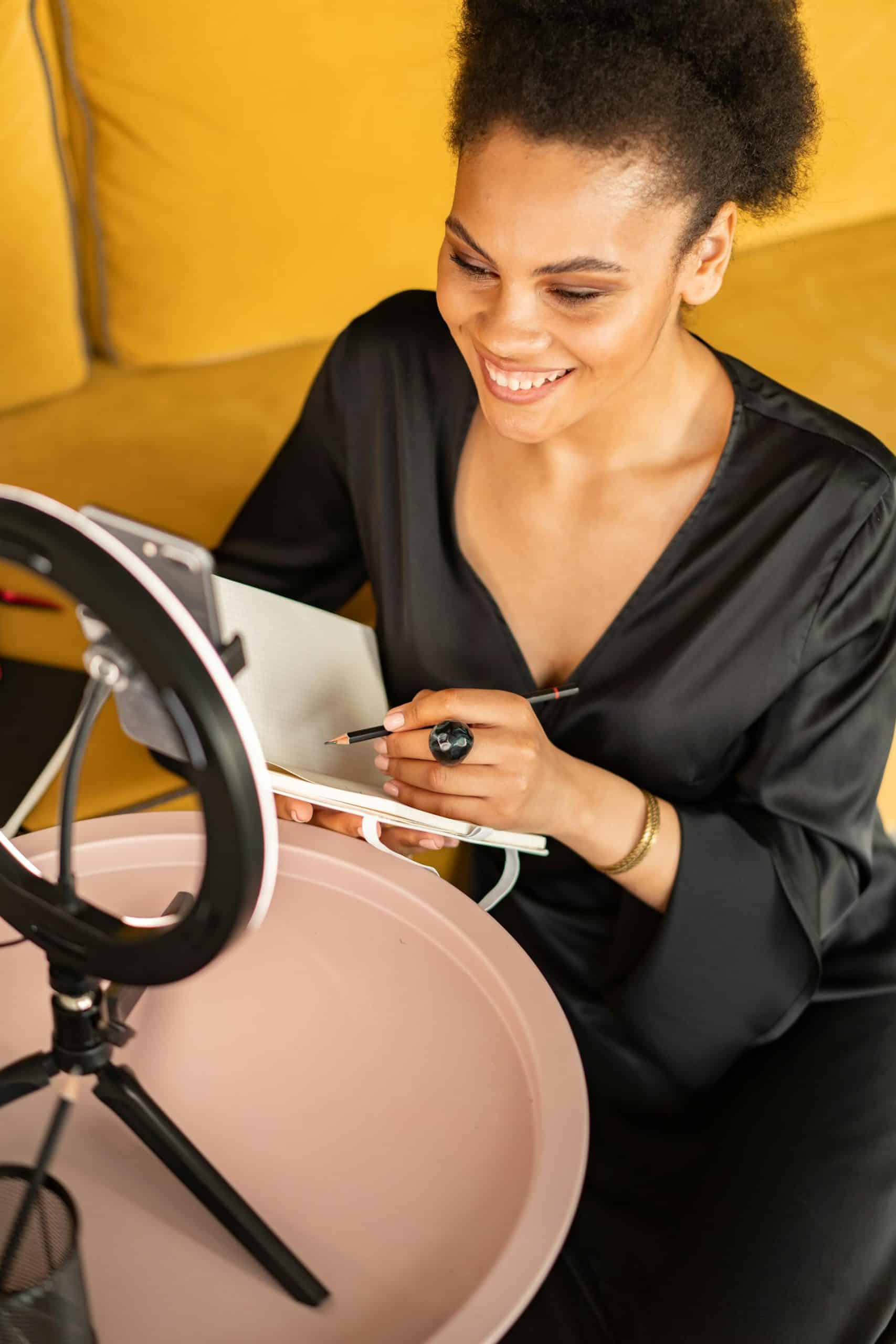 Young woman with afro joyfully creating on yellow sofa, illuminated by a ring light.