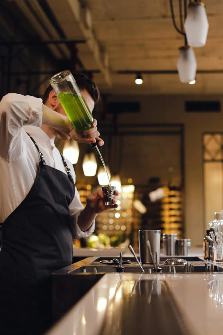 Skilled bartender pouring vibrant green cocktail in a chic bar with warm ambiance.
