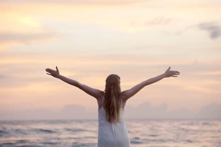 Woman joyfully embraces sunset at the beach, radiating freedom in a serene ocean setting.
