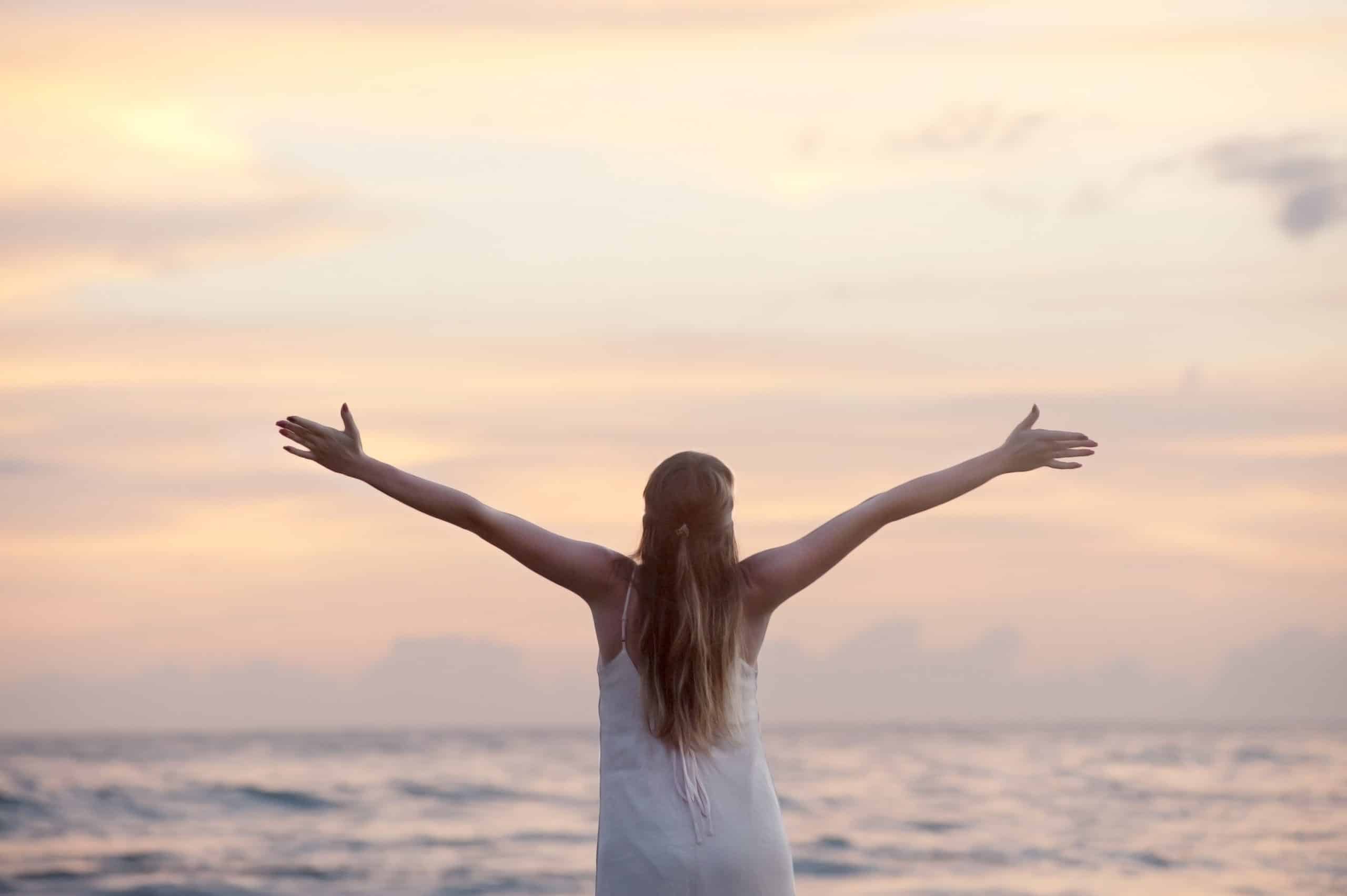 Woman joyfully embraces sunset at the beach, radiating freedom in a serene ocean setting.
