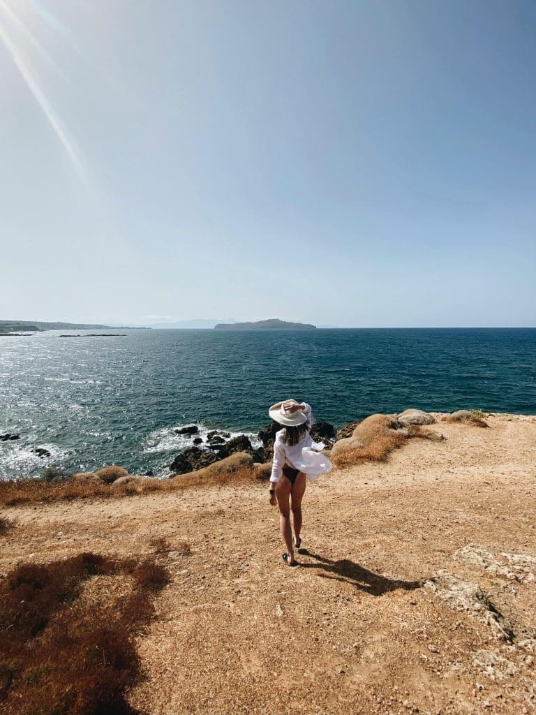 Woman in flowing attire gazes at tranquil blue sea, embodying peace and adventure by the coast.
