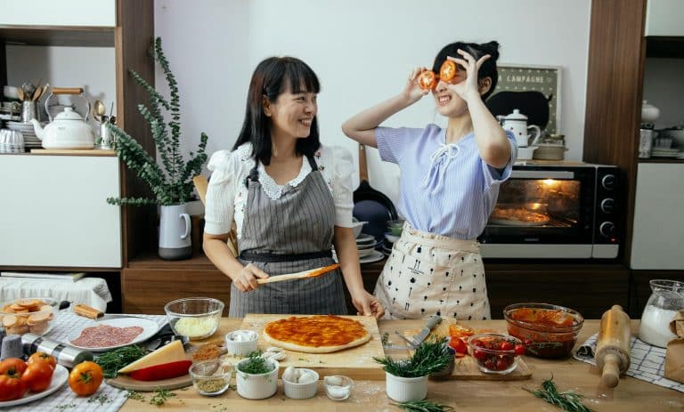 Two friends joyfully make pizza together in a bright, inviting kitchen filled with fresh ingredients.
