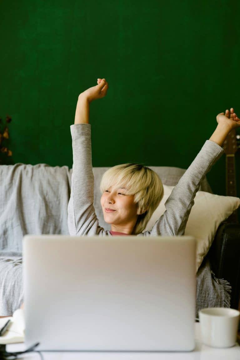 Young person relaxing on a couch, stretching with a laptop and cup in a cozy home.