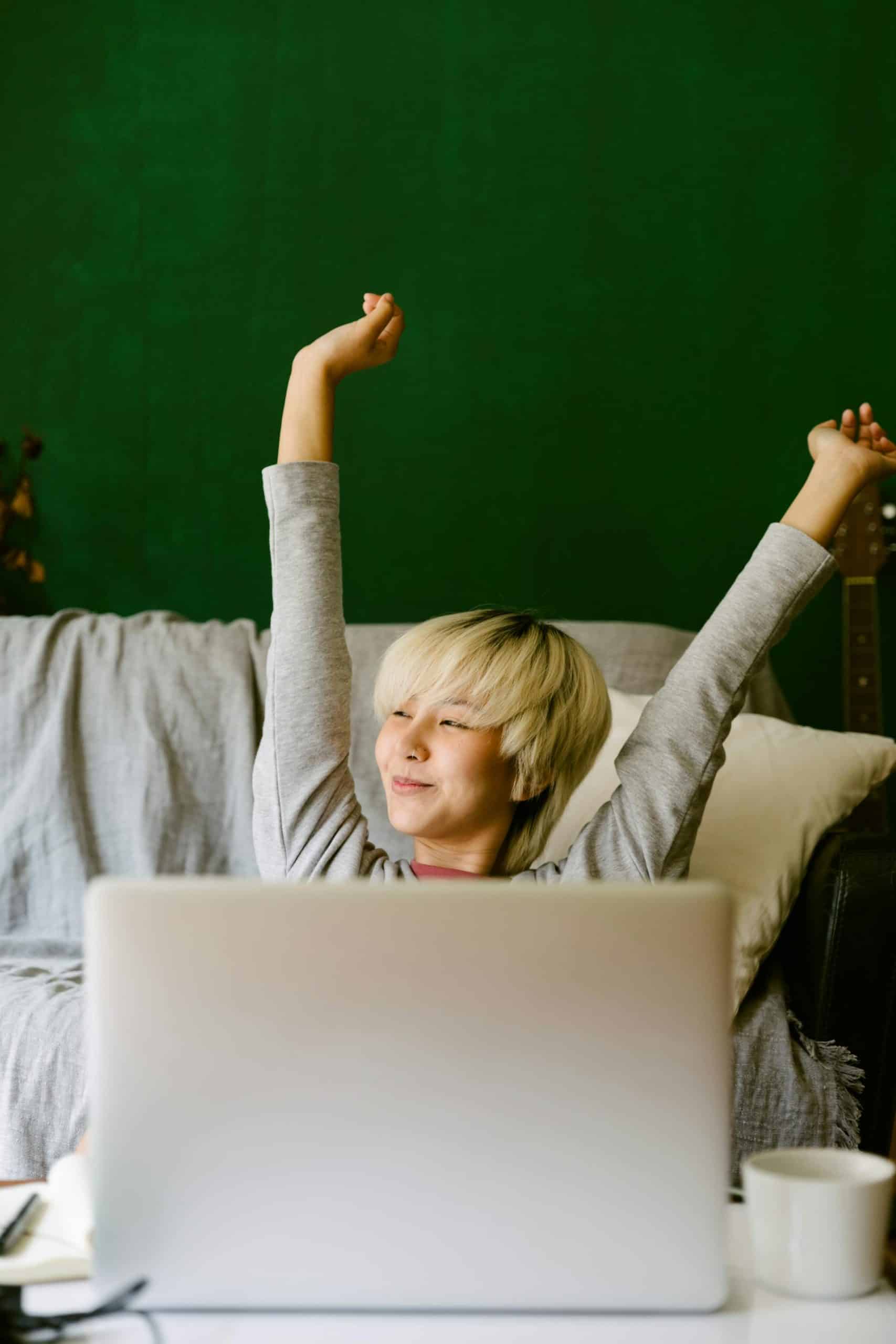 Young person relaxing on a couch, stretching with a laptop and cup in a cozy home.