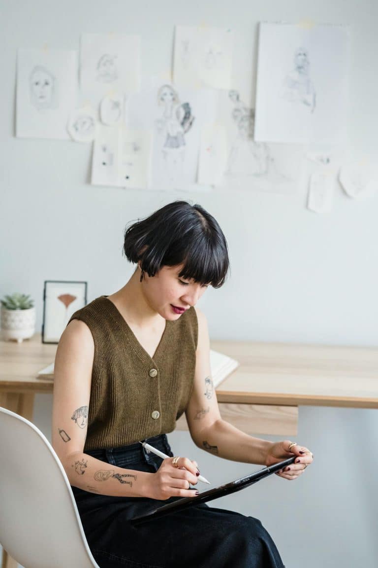 Woman in olive top focused on sketching at a minimalist desk surrounded by inspiring artwork.