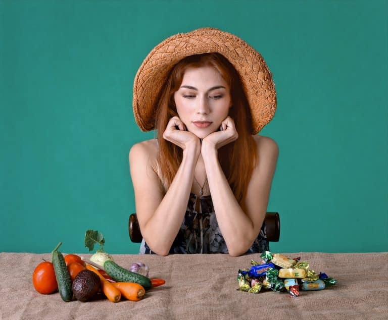 Young woman with red hair ponders healthy vegetables versus tempting candies in vibrant teal setting.