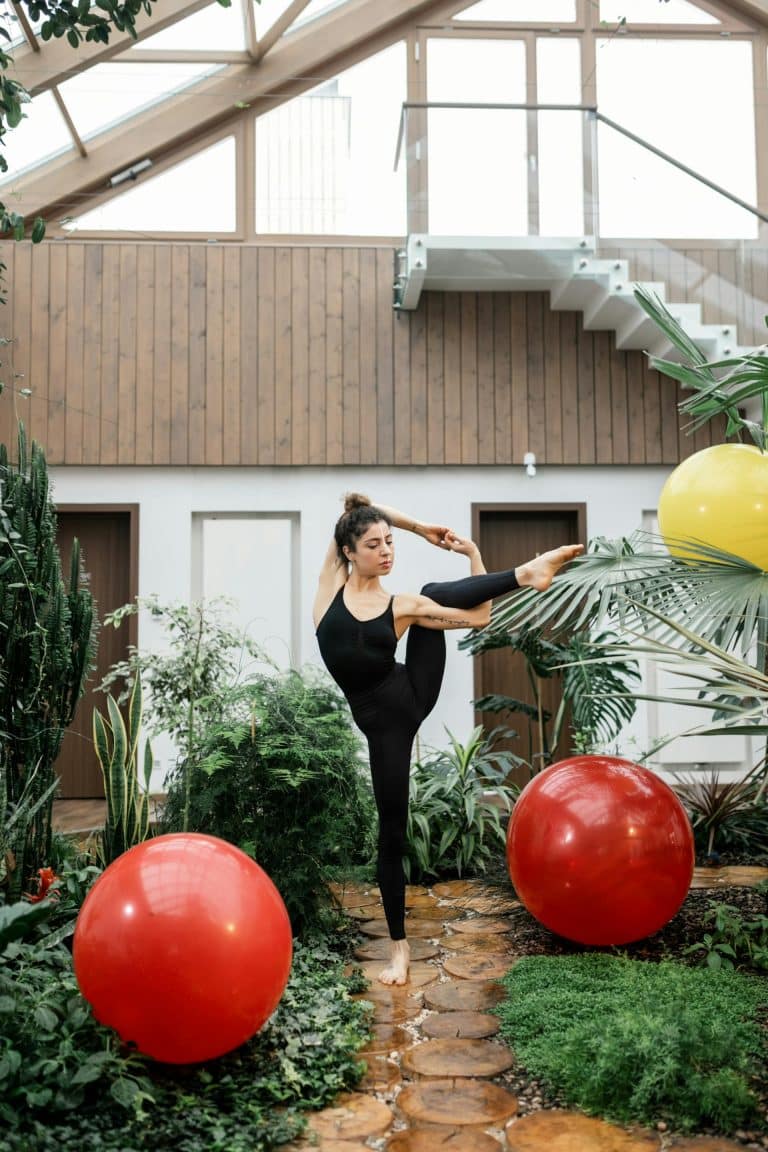 Elegant dancer in black bodysuit poses amid vibrant indoor garden with colorful spheres.