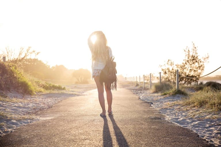 Woman walking barefoot on a coastal pathway at dusk, bathed in golden sunlight.