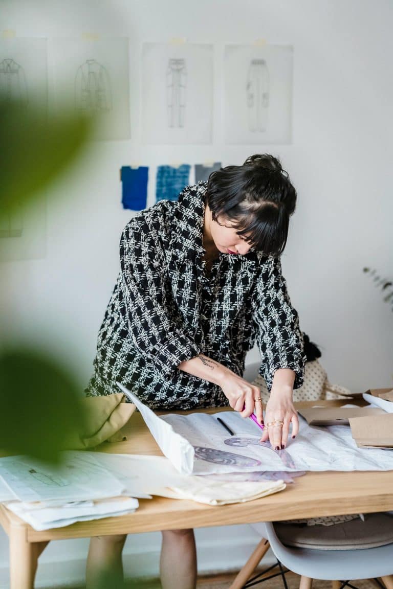 Fashion designer focused on sewing at a wooden table, surrounded by vibrant fabrics and sketches.
