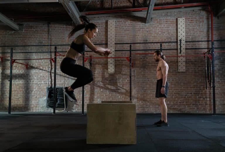 Woman jumps onto a box in a supportive gym environment, showcasing athleticism and teamwork.