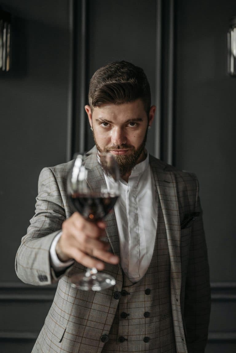Sophisticated man toasting with red wine in stylish gray plaid suit against a dark backdrop.