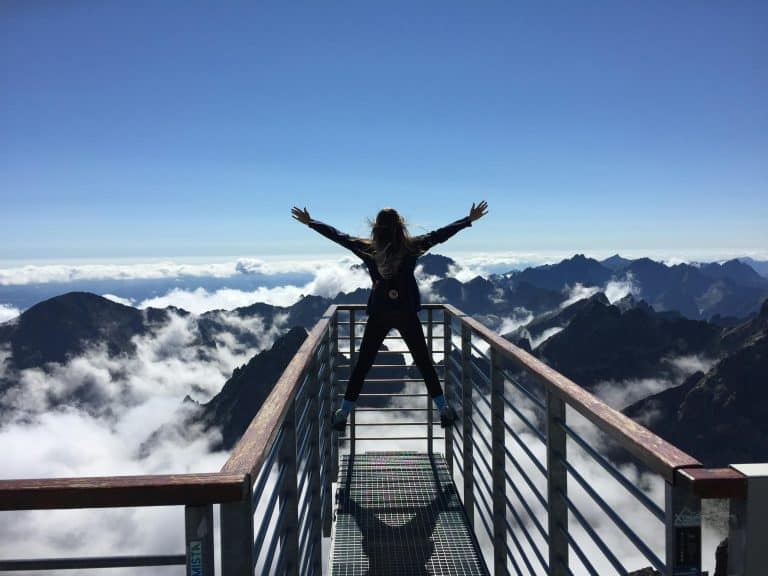 Person celebrating atop a mountain, arms outstretched above clouds and dramatic peaks.