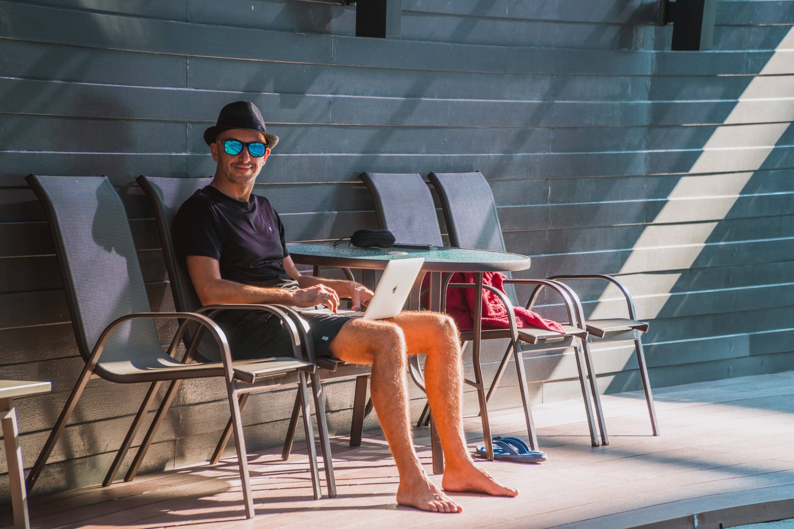 Man enjoys a relaxed day by the pool, working on his laptop in stylish summer attire.