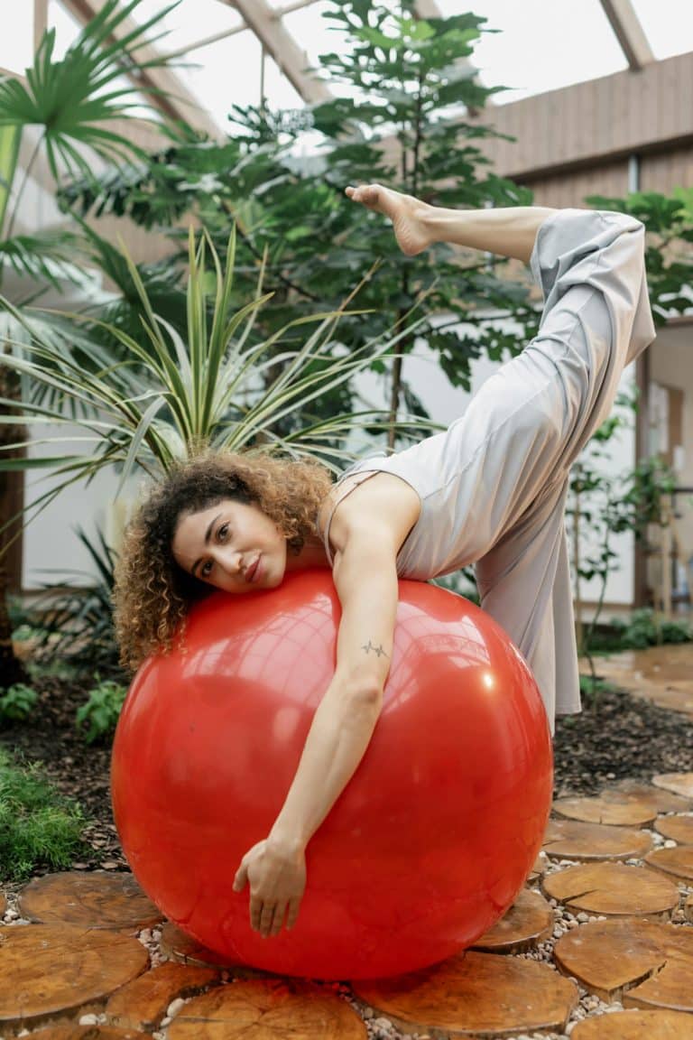 Young woman gracefully poses on a red ball amidst lush greenery and warm natural light.