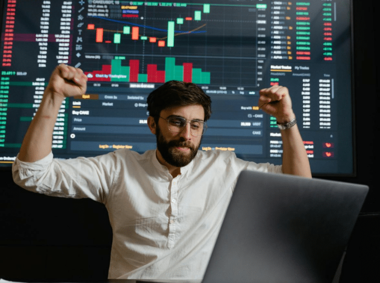 Young trader celebrates success at his desk with vibrant stock market charts in the background.