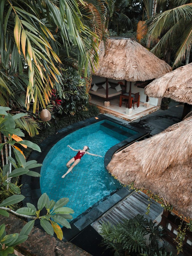 Tranquil tropical pool scene with floating figure, lush greenery, and sunlit thatched roofs.