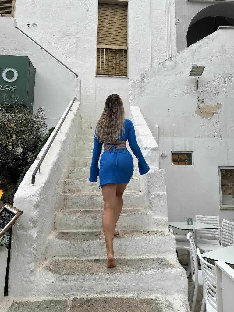 Woman in blue dress gracefully climbs weathered white stairs in a charming outdoor setting.