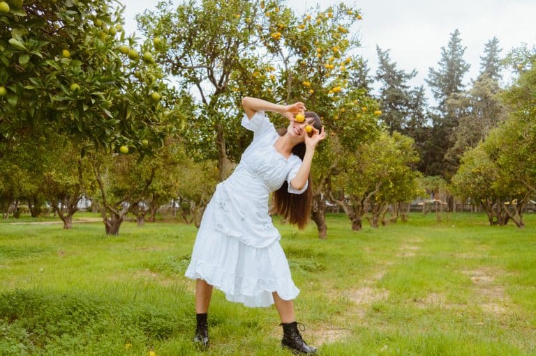 Woman in a white dress joyfully poses with orange fruits in a vibrant orchard.