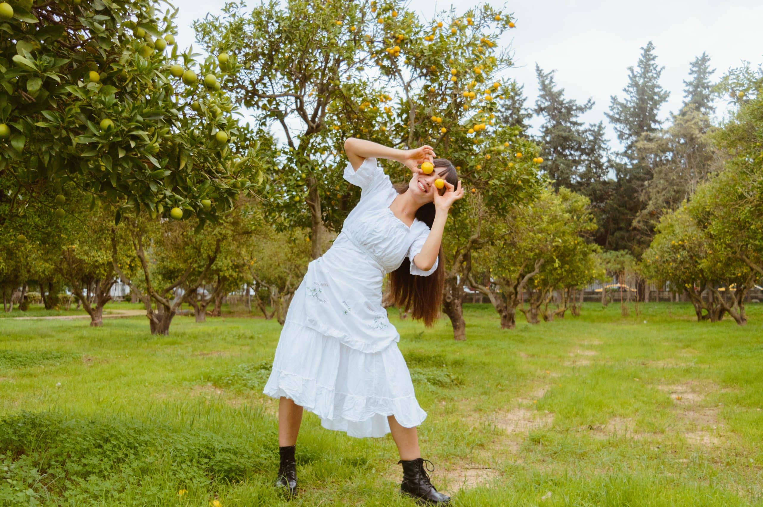 Woman in a white dress joyfully poses with orange fruits in a vibrant orchard.