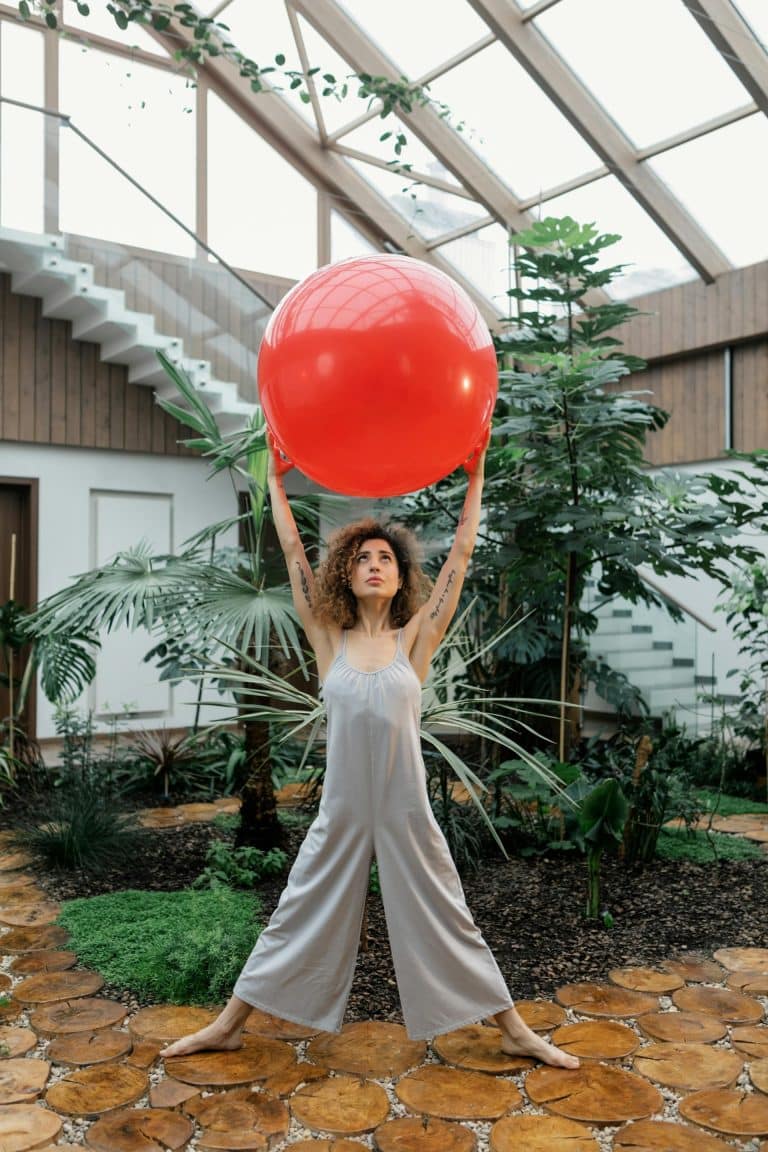 Confident woman lifts red exercise ball in a bright, green indoor fitness oasis.