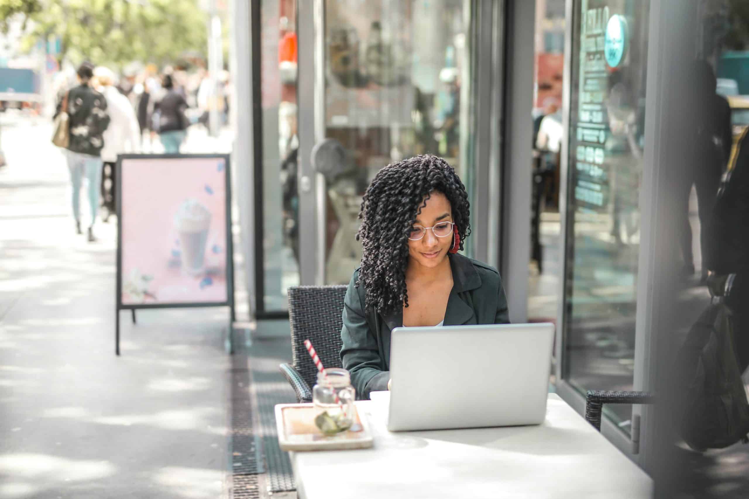 Woman with curly hair works on a laptop at a lively outdoor cafÃ©.