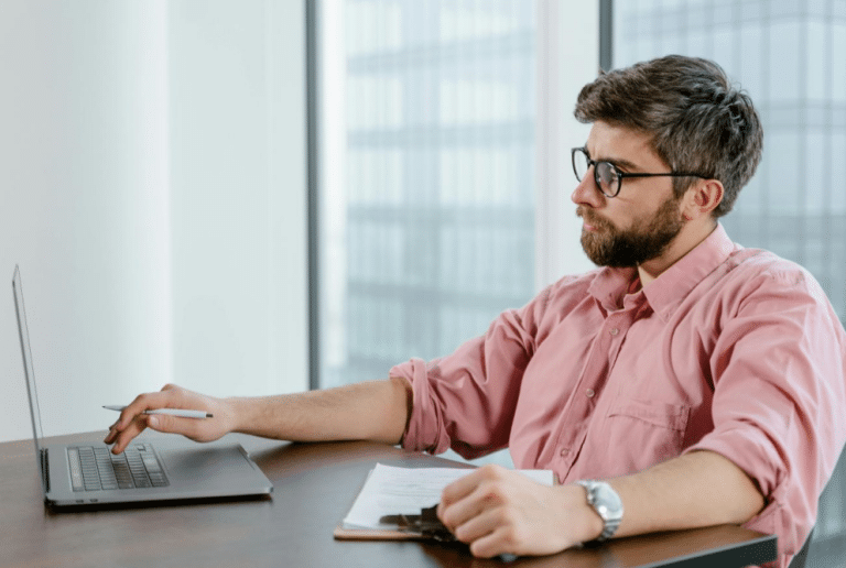 Young man in light pink shirt focused on laptop in bright modern office space.