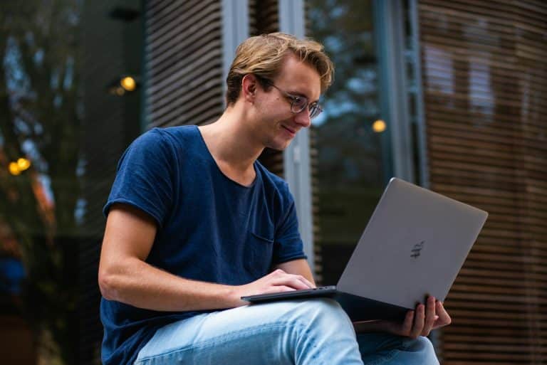 Young man working on laptop outdoors, enjoying a relaxed atmosphere with greenery and modern architecture.
