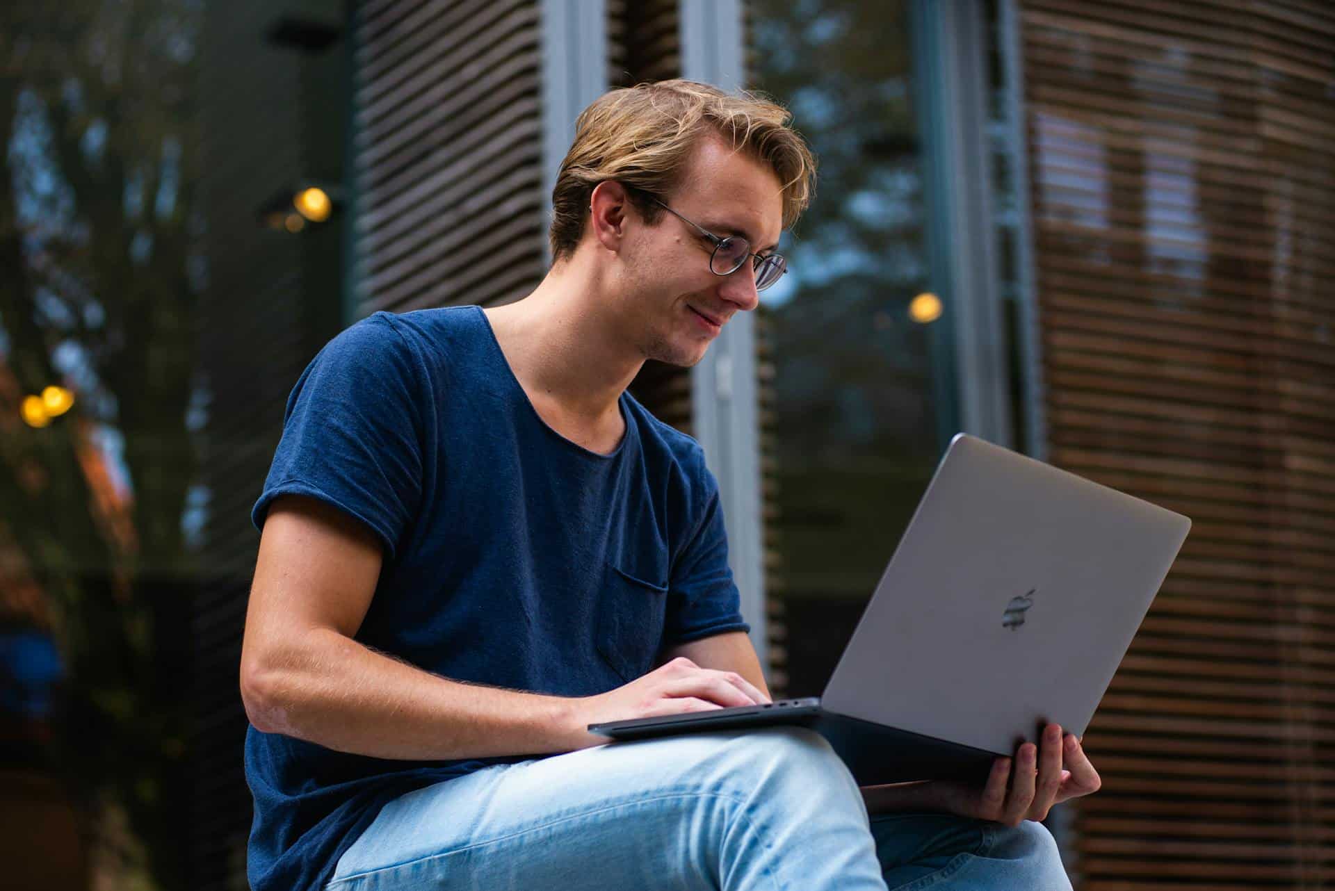 Young man working on laptop outdoors, enjoying a relaxed atmosphere with greenery and modern architecture.