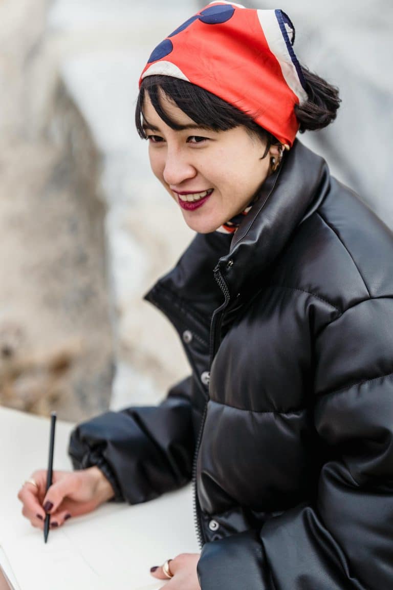 Cheerful young woman artist drawing outdoors in stylish jacket and vibrant scarf against nature.