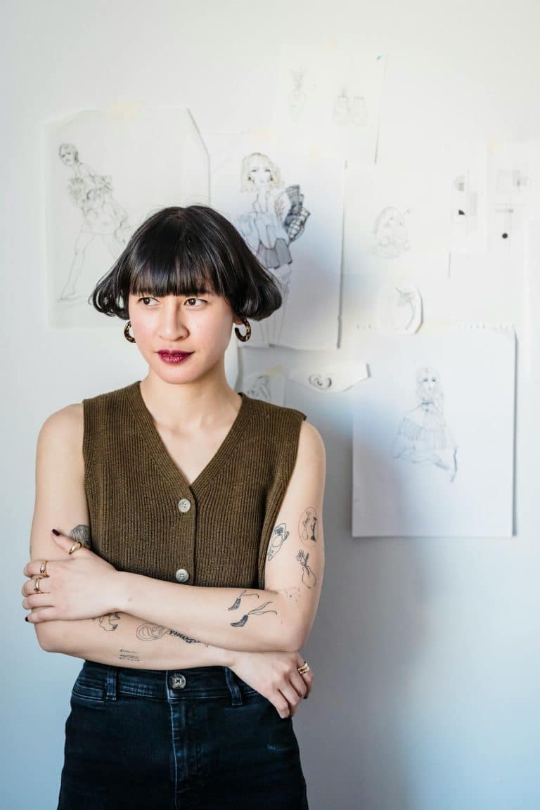 Confident woman with tattoos in artistic space, wearing olive top and bold lipstick.