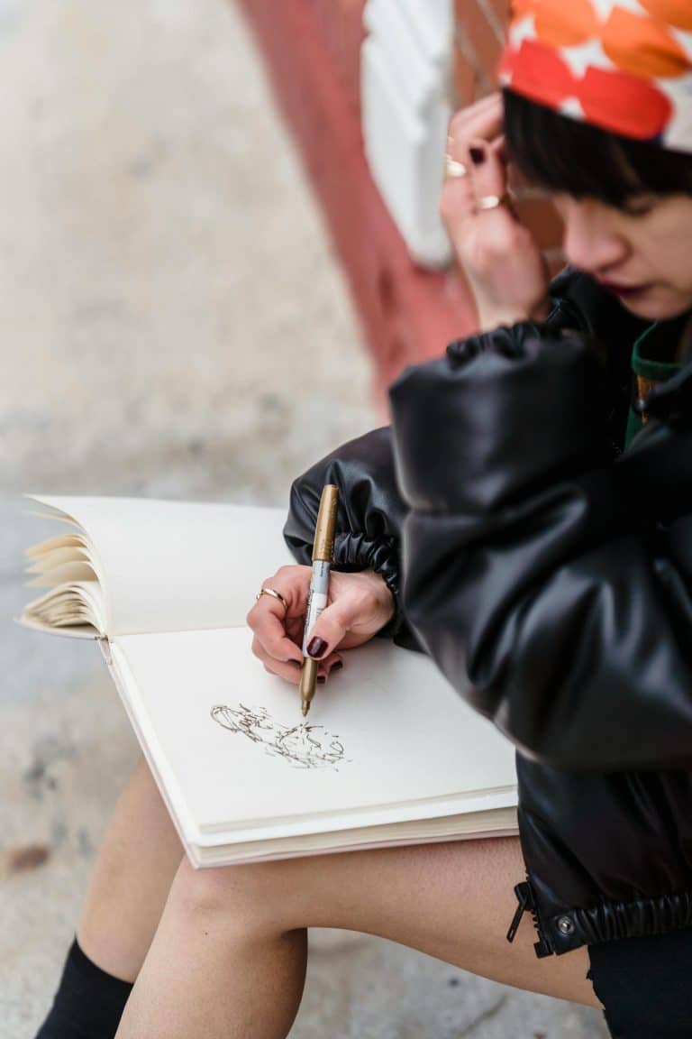 Young woman sketching in an urban setting, wearing a black jacket and colorful headscarf.