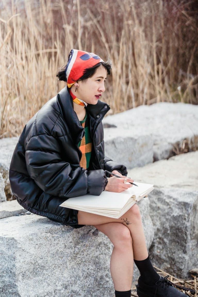 Young woman sketches in nature, wearing a stylish puffer jacket, colorful sweater, and vibrant scarf.