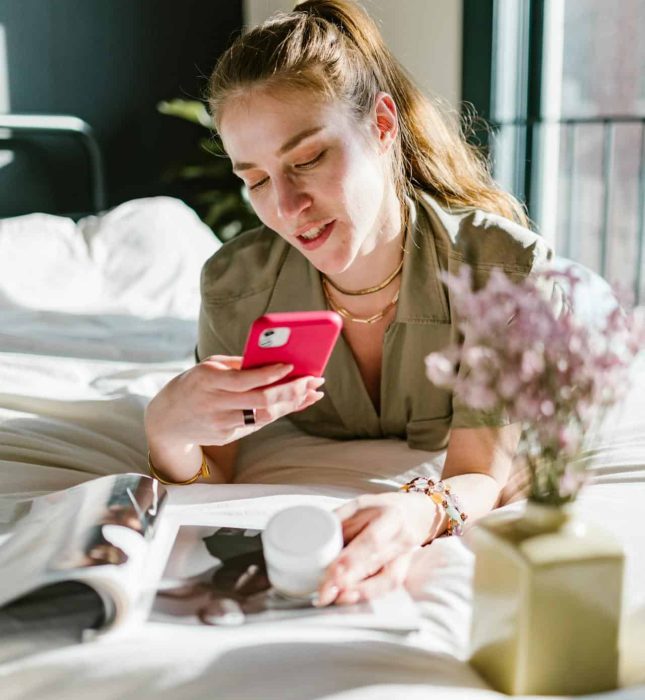 Young woman enjoying cozy self-care moment on bed with sunlight, smartphone, magazine, and flowers.