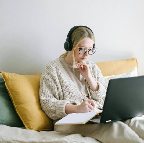 Young woman wearing a cardigan works on her laptop in a cozy, inviting bedroom.