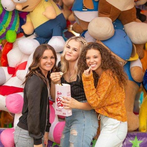 Three friends joyfully pose with oversized stuffed animals at a colorful carnival.