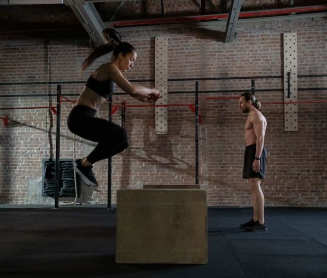 Woman jumps onto a box in a supportive gym environment, showcasing athleticism and teamwork.