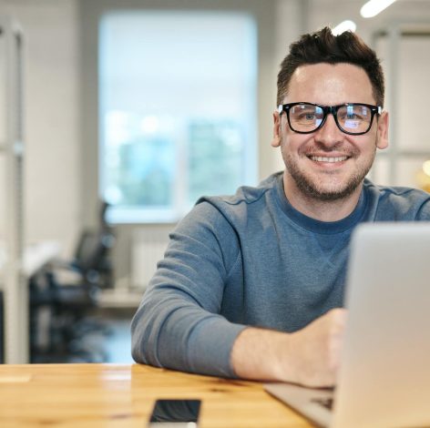 Cheerful young man in a grey sweater working on a laptop in a bright office.