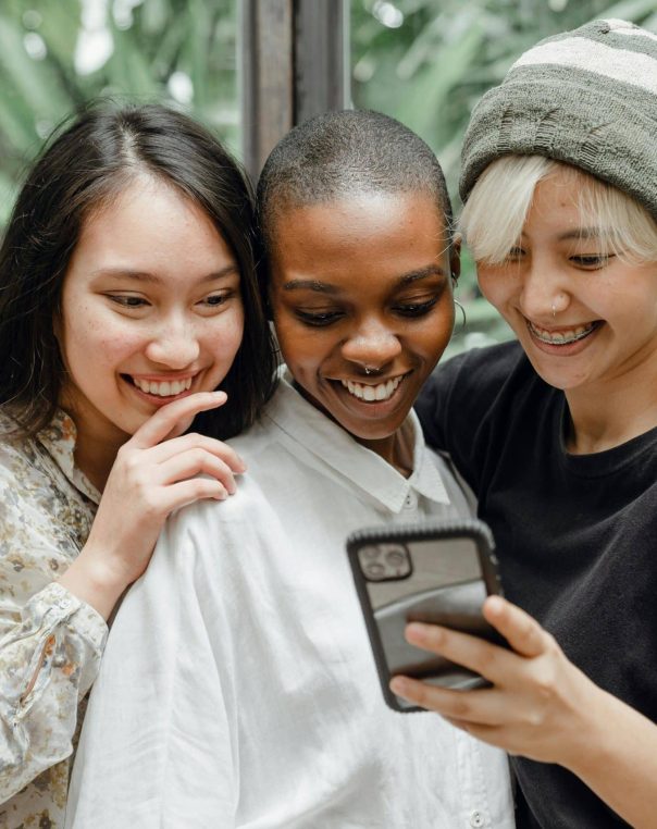 Three joyful friends share laughter while looking at a smartphone indoors, surrounded by greenery.