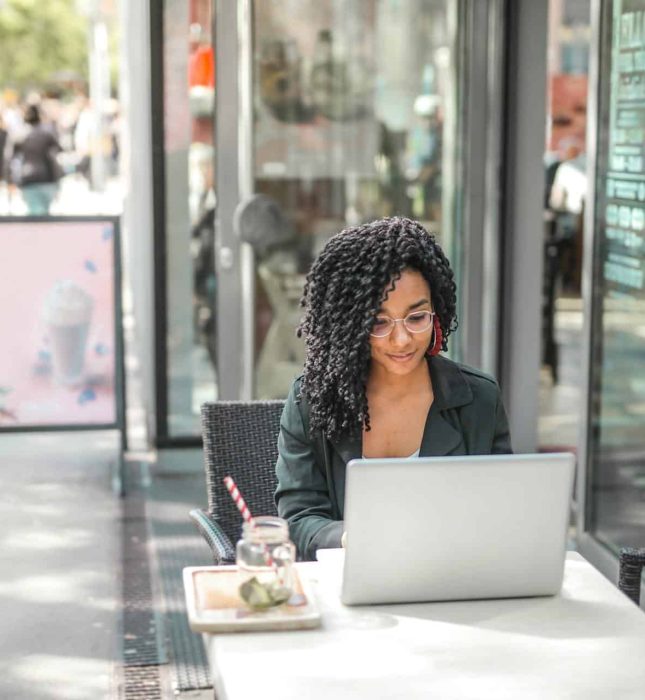 Woman working on laptop at a lively outdoor cafÃ©, enjoying a snack and drink.