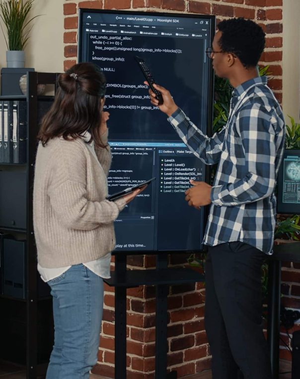 Team of database admins analyzing source code on wall screen tv comparing errors using digital tablet in busy server room. Two cloud programers debugging algorithm in software innovation office.