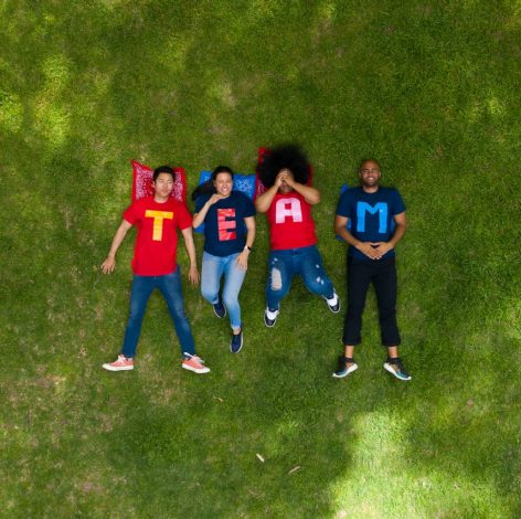 Four friends spell TEAM on lush grass, showcasing teamwork and joy in colorful t-shirts.