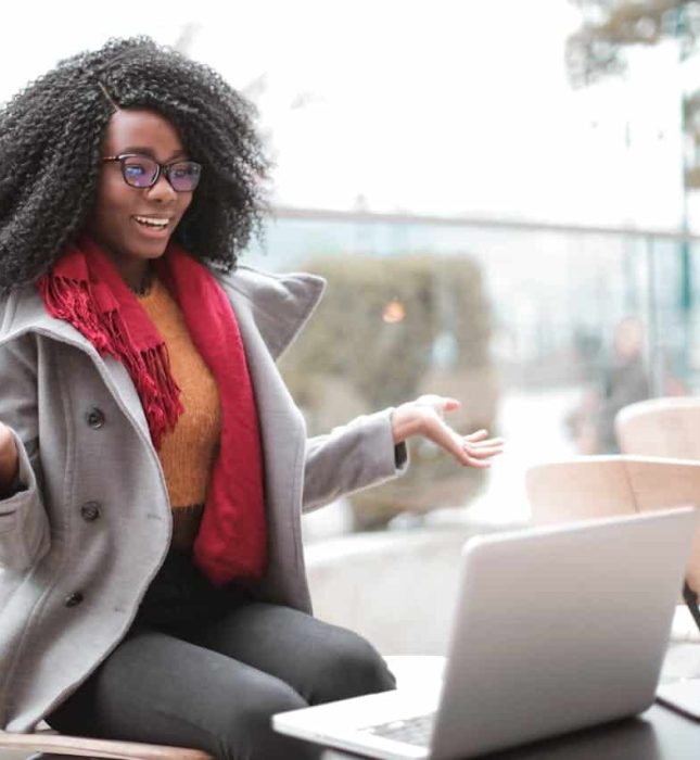 Young woman in gray coat and red scarf enthusiastically video calling at outdoor cafÃ©.