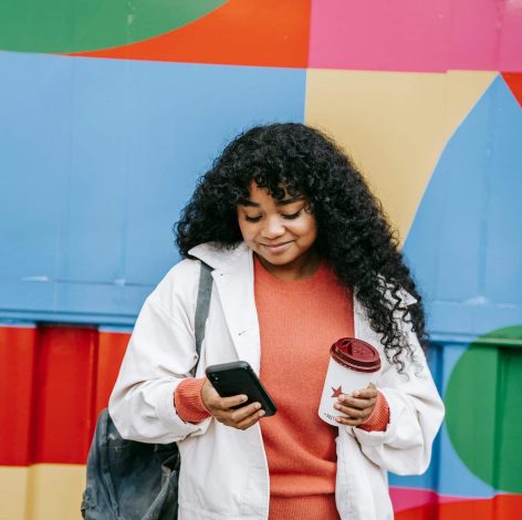 Casual young woman interacts with smartphone against a vibrant geometric wall, enjoying her drink.