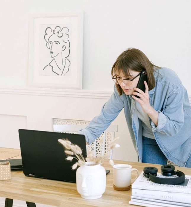 Focused woman multitasking at a stylish home office desk with laptop and smartphone.