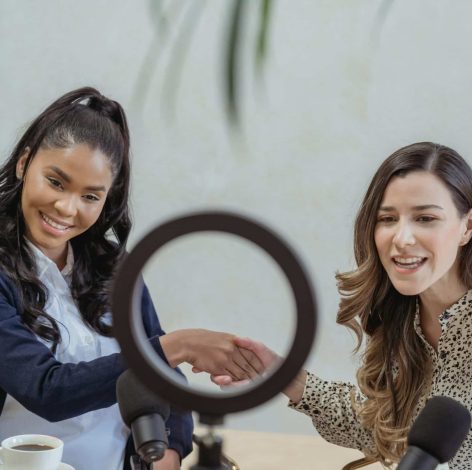 Two women enjoy a lively podcast conversation, illuminated by a ring light and microphones.