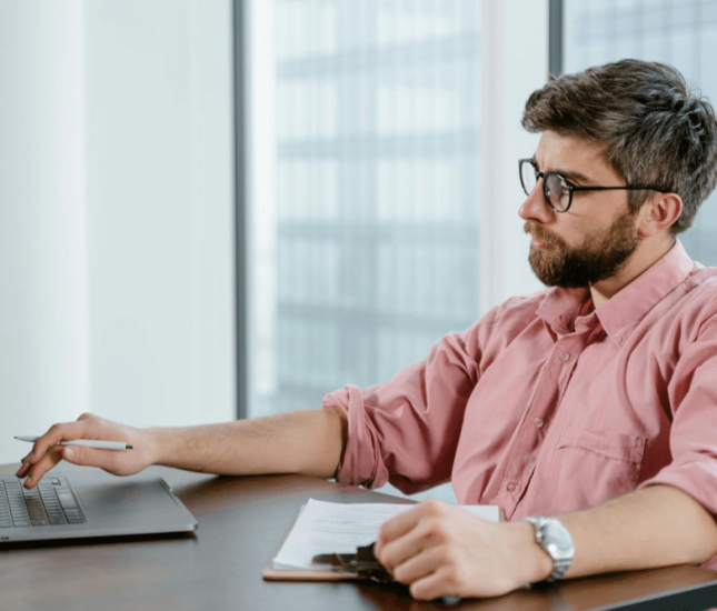 Young man in light pink shirt focused on laptop in bright modern office space.