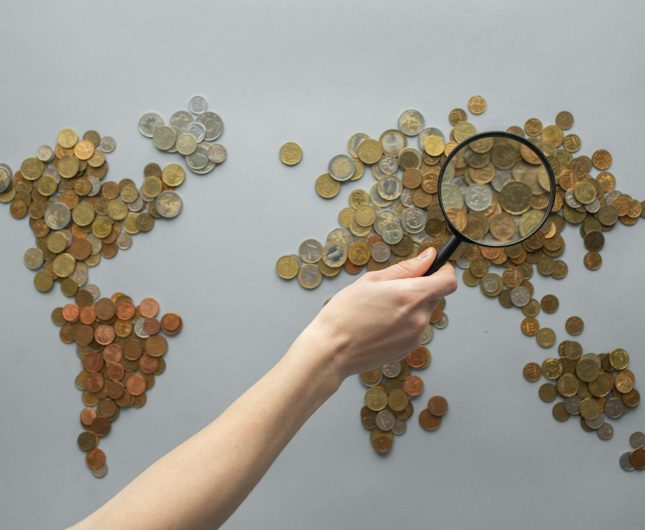 World map made of coins, highlighting global currencies and economic connections through a magnifying glass.
